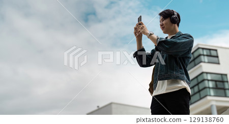 Young Man Enjoying Music and Photography Outdoors with Smartphone and Headphones Against Modern Urban Background Young Man Enjoying Music and Photography Outdoors with Smartphone and Headphones Against Modern Urban Background 129138760