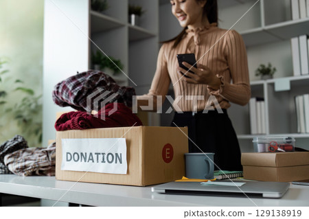 Woman Organizing Clothes for Donation in a Cardboard Box at Home Office with Smartphone and Office Supplies Woman Organizing Clothes for Donation in a Cardboard Box at Home Office with Smartphone and Office Supplies 129138919