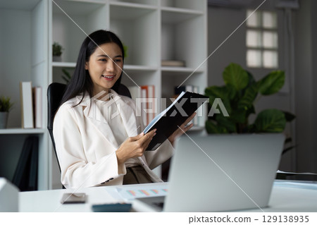 Confident Businesswoman Analyzing Documents in Modern Office Setting with Laptop and Greenery 129138935