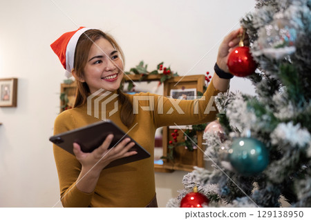 Woman Decorating Christmas Tree with Ornament and Tablet in Hand, Wearing Santa Hat, Celebrating Holiday Season Indoors Woman Decorating Christmas Tree with Ornament and Tablet in Hand, Wearing Santa Hat, Celebrating Holiday Season Indoors 129138950