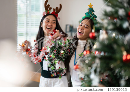 Festive Celebration with Friends Wearing Christmas Accessories and Decorating a Christmas Tree Indoors Festive Celebration with Friends Wearing Christmas Accessories and Decorating a Christmas Tree Indoors 129139013