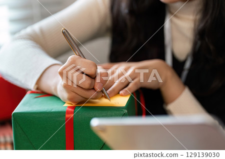 Woman Writing on Gift Tag Attached to Green Wrapped Present with Red Ribbon During Holiday Season 129139030