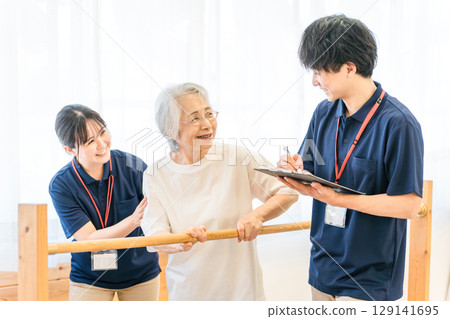 Senior woman using handrails for walking training and male and female occupational therapist and physiotherapist staff assisting her 129141695