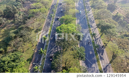 An aerial view of a wide, multi-lane road flanked by trees, with cars and motorbikes driving on a sunny day. An aerial view of a wide, multi-lane road flanked by trees, with cars and motorbikes driving on a sunny day. 129145954