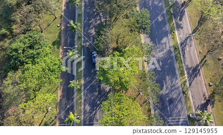 An aerial view of a wide, multi-lane road flanked by trees, with cars and motorbikes driving on a sunny day. An aerial view of a wide, multi-lane road flanked by trees, with cars and motorbikes driving on a sunny day. 129145955