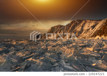 Beautiful winter landscape of frozen Lake Baikal at sunrise - A granite rock with steep slopes rises above a frozen lake. Winter ice covers providing a stunning backdrop for winter tourism adventures. Beautiful winter landscape of frozen Lake Baikal at sunrise - A granite rock with steep slopes rises above a frozen lake. Winter ice covers providing a stunning backdrop for winter tourism adventures. 129147221