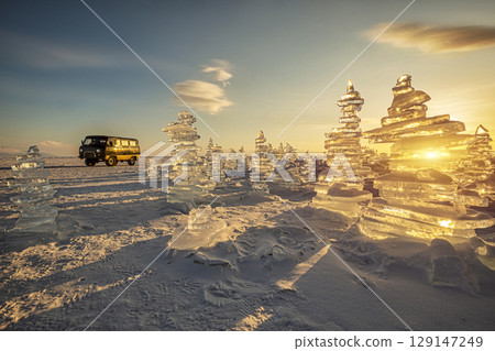 Sunset reflected in a large block of ice hummock and a classic vintage van car on the frozen lake background. 129147249