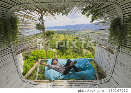Asian man lying down on sofa bed Asian tourist man is lying down on sofa bed near terrace in cafe outdoor with scenic view on mountains. Time to relax Asian man lying down on sofa bed Asian tourist man is lying down on sofa bed near terrace in cafe outdoor with scenic view on mountains. Time to relax 129147912
