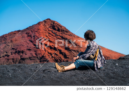 A girl sits on a black sand, enjoys a volcanic mountain landscape. 129148662