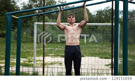Young muscular sportsman doing pull ups at sports ground. Athletic guy exercising outdoor. Strong sporty man working out at summer nature. Concept of healthy and active lifestyle. Slow motion Young muscular sportsman doing pull ups at sports ground. Athletic guy exercising outdoor. Strong sporty man working out at summer nature. Concept of healthy and active lifestyle. Slow motion 129151659