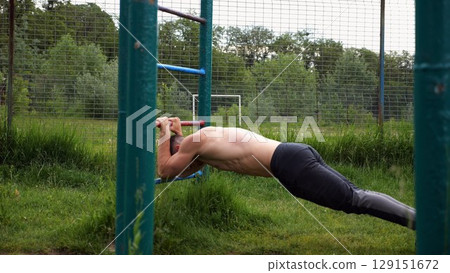 Young muscular athlete doing exercise on triceps at sport equipment outdoor. Male shirtless athlete working out at sport ground. Strong sporty man training at nature. Concept of healthy lifestyle Young muscular athlete doing exercise on triceps at sport equipment outdoor. Male shirtless athlete working out at sport ground. Strong sporty man training at nature. Concept of healthy lifestyle 129151672