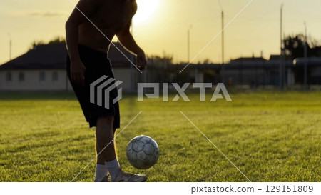 Male feet of professional footballer kicking ball at green field. Legs of young man juggling soccer ball on stadium at sunset. Sportsman practicing tricks outdoor. Freestyle football concept 129151809