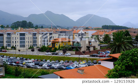 Cityscape of a Llanes, Spain with a mix of modern and traditional buildings, a crowded parking lot, and majestic mountains in the background under a cloudy sky Cityscape of a Llanes, Spain with a mix of modern and traditional buildings, a crowded parking lot, and majestic mountains in the background under a cloudy sky 129153926