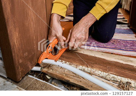 Plumber uses pipe cutter to remove leaking section from plastic tubing during replacement process at area where visible damage occurred earlier. Plumber uses pipe cutter to remove leaking section from plastic tubing during replacement process at area where visible damage occurred earlier. 129157127