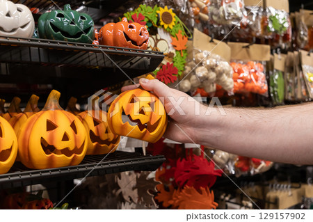Male hand selecting a classic orange jack-o'-lantern decoration from a crowded retail store shelf, highlighting popular Halloween themed products for festive autumn shopping Male hand selecting a classic orange jack-o'-lantern decoration from a crowded retail store shelf, highlighting popular Halloween themed products for festive autumn shopping 129157902