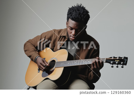 A young man playing an acoustic guitar, seated casually on a stool against a neutral gray background 129160360