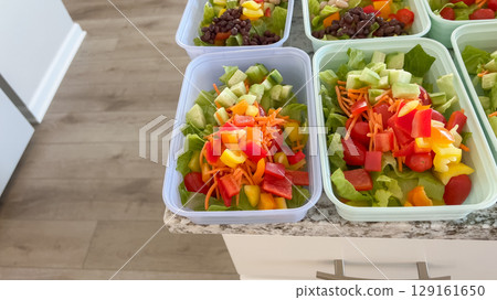 Rows of organized plastic salad containers are filled with chopped vegetables and leafy greens, arranged for weekly meal prep in a bright kitchen setting. Rows of organized plastic salad containers are filled with chopped vegetables and leafy greens, arranged for weekly meal prep in a bright kitchen setting. 129161650