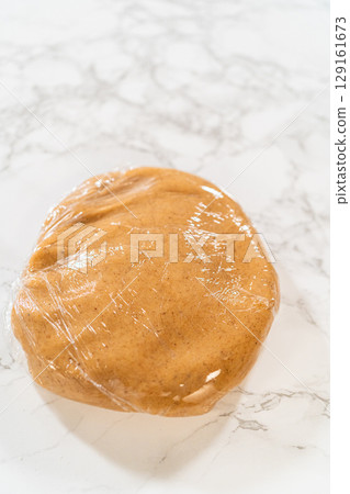 Close-up view of the Amber Gingerbread Cookie Dough wrapped in plastic wrap, shaped into a flat disc for even chilling in the refrigerator. The wrapping preserves freshness and prepares the dough for 129161673