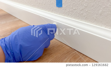 Close-up of a hand in a blue glove inspecting or cleaning a white baseboard trim set against a tan wall and wood-style flooring. The image is well-lit and focused on home improvement or maintenance 129161682