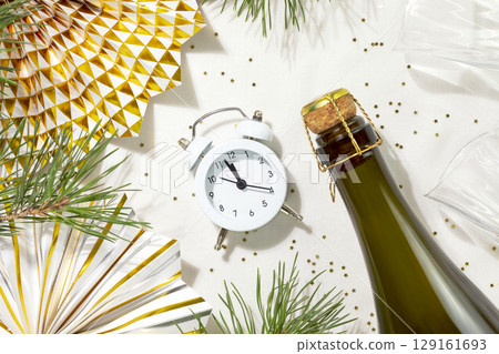 Winter holidays card with fir branches, bottle of champaign, wine glasses, clock and holiday decoration on a white table with golden confetti. Christmas and New Year flat lay 129161693