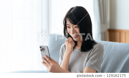Smiling woman sitting on sofa and using smartphone 129162175