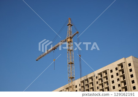 A majestic construction crane stands impressively tall against a vibrant and clear blue sky above A majestic construction crane stands impressively tall against a vibrant and clear blue sky above 129162191