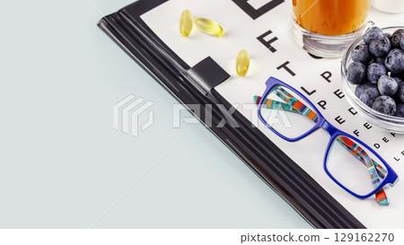 Kid eyeglasses, glass of carrot juice, bowl with blueberries and Snellen table for vision testing on blue background with copy space. Nutritions for good vision. Vitamin A product to improve vision 129162270