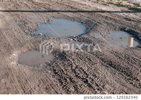 Close-up of a dirt road with several puddles of water, showcasing the texture of the ground and the reflections. Close-up of a dirt road with several puddles of water, showcasing the texture of the ground and the reflections. 129162340