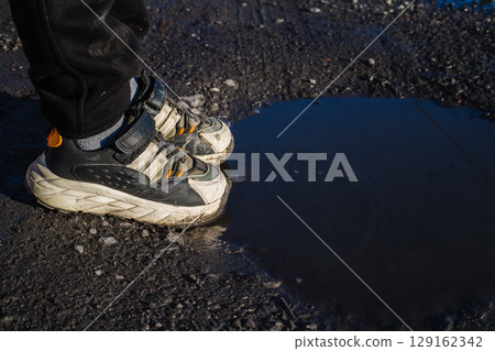 Close-up of children's feet in sneakers standing on a dirt road near a puddle Close-up of children's feet in sneakers standing on a dirt road near a puddle 129162342