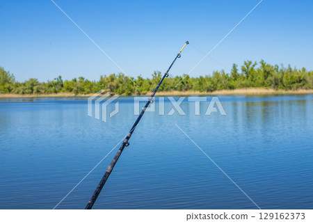 Fishing rod extends towards the water on a sunny day, capturing a beautiful outdoor scene 129162373