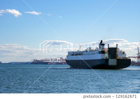 Under a clear blue sky, a large cargo ship sails across the vast ocean, and a port crane can be seen in the distance. 129162671