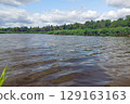 A wide river with dark water and green thickets along the banks under a cloudy summer sky. 129163163