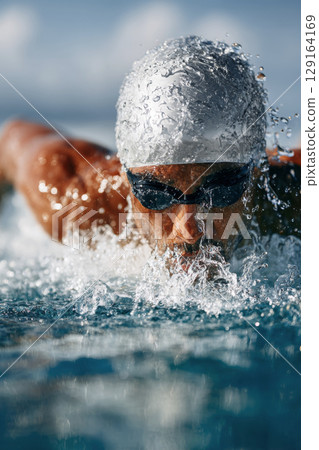 Competitive swimmer in silver cap and goggles performing a powerful stroke in clear water Competitive swimmer in silver cap and goggles performing a powerful stroke in clear water 129164169