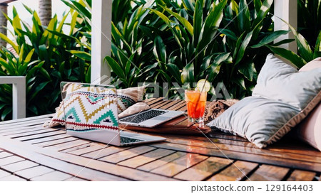 Laptop resting beside tropical cocktail on wooden surface, plush pillows framing relaxed workspace surrounded by verdant landscape, signaling digital nomad lifestyle 129164403