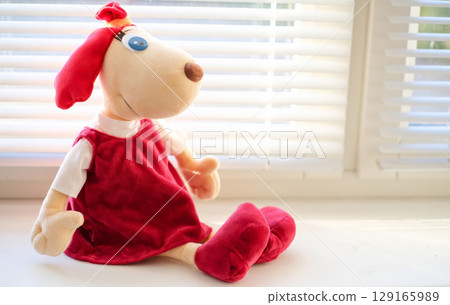 A cute plush dog toy Lotte dressed in a red dress sits on a sunlit windowsill, looking out the window with a gentle expression 129165989