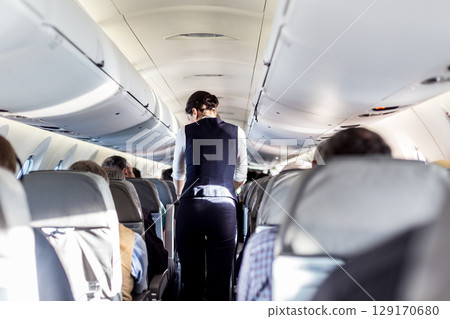 Interior of airplane with passengers on seats and stewardess in uniform walking the aisle, serving people. Commercial economy flight service concept 129170680