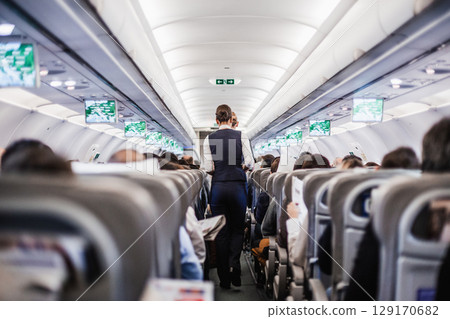 Interior of airplane with passengers on seats and stewardess in uniform walking the aisle, serving people. Commercial economy flight service concept 129170682
