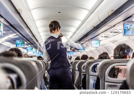 Interior of airplane with passengers on seats and stewardess in uniform walking the aisle, serving people. Commercial economy flight service concept Interior of airplane with passengers on seats and stewardess in uniform walking the aisle, serving people. Commercial economy flight service concept 129170683
