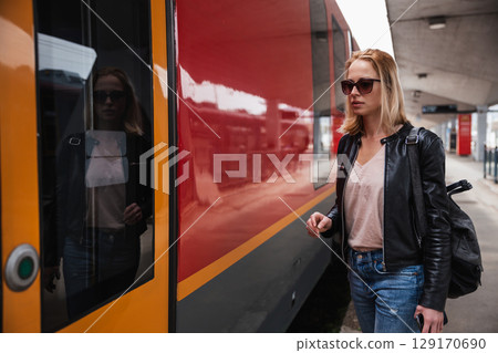 Young blond woman in jeans, shirt and leather jacket wearing bag and sunglass, embarking red modern speed train on train station platform. Travel and transportation Young blond woman in jeans, shirt and leather jacket wearing bag and sunglass, embarking red modern speed train on train station platform. Travel and transportation 129170690