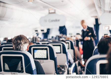 Interior of airplane with passengers on seats and stewardess in uniform walking the aisle, serving people. Commercial economy flight service concept 129170801