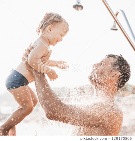 Dad and baby under a tropical shower. dad and his cute infant boy playing on the beach under shower water splashes - Father taking care of cute sun in playful moment at summer family holiday. 129170806