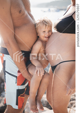 Funny summer family portrait of cute little smiling boy with his head squeezed between two large bellies of his parents who expect another baby. 129170807
