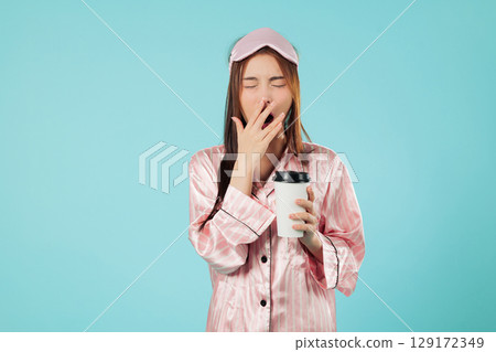 Tired young woman in pajamas and sleep mask holding coffee cup, needs that morning coffee to wake up as she rubs her eye while resting at home, headshots isolated studio blue background 129172349