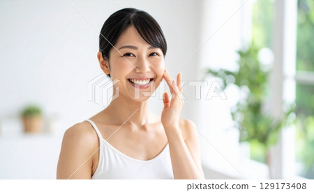 A smiling young woman putting her hands on her cheeks in front of a mirror to do skin care, in a bright room 129173408
