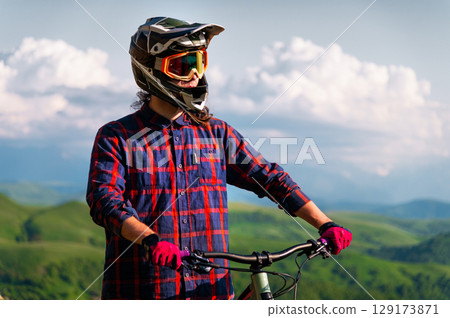 Portrait of a cyclist standing with his bicycle against a dramatic sky with cumulus clouds. Handsome young man biking in the countryside 129173871