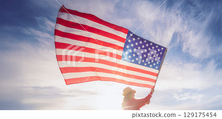 Close up on USA flag in a hand of a person against the sky. Independence Day or travelling in America concept. Panorama of the American flag against the blue sky 129175454