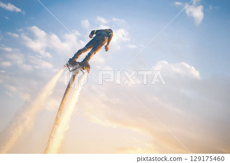 A flyboard athlete hovers over the water against the sunset sky, propelled by powerful jet streams of water. Flyboarding adventure at sunset over calm waters showcasing an exhilarating water sport 129175460