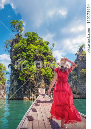 A Woman female solo traveler in a bright red dress and a wide-brimmed straw hat Standing on a Traditional Thai Longtail Boat and looking at the broken rocks on Cheo Lan Lake in Khao Sok Park in 129175464