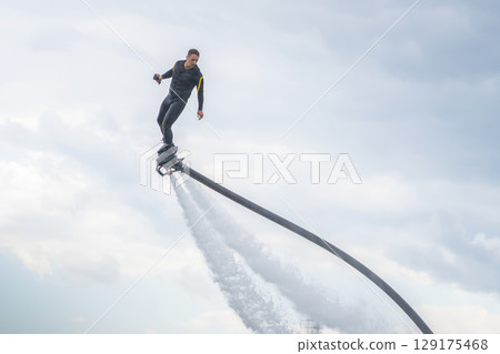 A man in a black athlete wetsuit hovers against a dramatic sky on a flyboard. A performer demonstrates impressive flyboarding skills, soaring above the clear blue water. Dressed in a sleek wetsuit, A man in a black athlete wetsuit hovers against a dramatic sky on a flyboard. A performer demonstrates impressive flyboarding skills, soaring above the clear blue water. Dressed in a sleek wetsuit, 129175468
