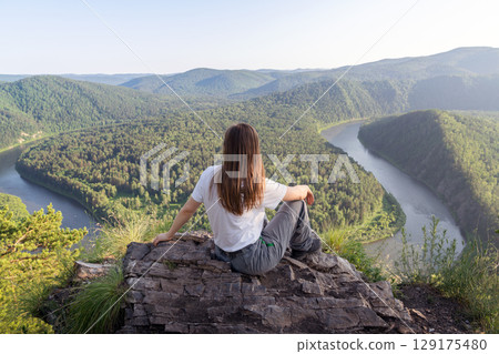 A person sits on a rocky ledge, gazing at a breathtaking view of a winding river framed by rolling hills and dense forests. A female solo traveler rests on a mountain and looks at the Mana River. The 129175480